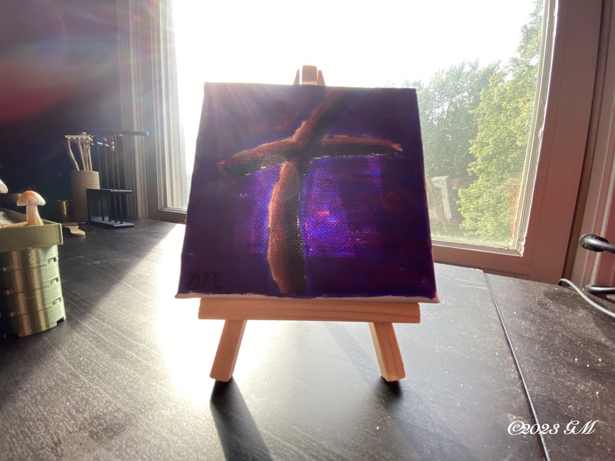 At the Cross painting on a mini easel being displayed on a desk in front of a window. Sunlight is coming in through the window to highlight the painting.